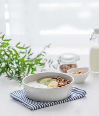 Muzlu ve kivili granola beyaz bir masada, pencerenin arka planında yeşil bir dal var. Sağlıklı kahvaltı konsepti vejetaryen yoğurt, müsli ve taze meyve..