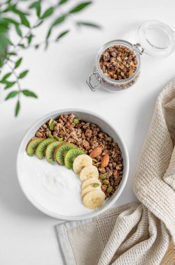 Yeşil dalı ve peçetesi olan beyaz bir masada muz ve kivi ile granola. Sağlıklı kahvaltı konsepti: Vejetaryen yoğurt, fındık, müsli ve taze meyve. Üst görünüm. 