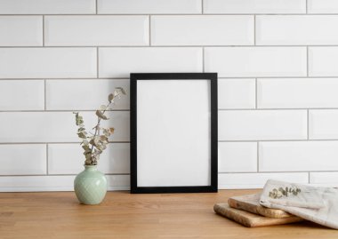 Beyaz kiremit bir duvara karşı boş bir tuval ve vazo ve kesme tahtasıyla ahşap bir masanın üzerinde siyah bir çerçeve. Fotoğraf, poster ya da resim tasarımı. Boşluğu kopyala.