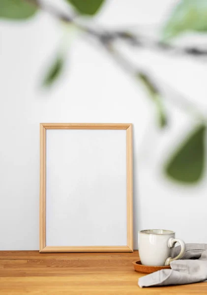 A  frame with a blank canvas against a light wall and on a wooden tabletop with blurred green foliage and cup of tea. Layout concept for photography, poster or painting. Copy space.