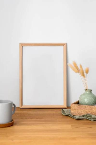 A frame with a blank canvas against a light wall and on a wooden tabletop with vase and cup of tea. Layout concept for photography, poster or painting. Copy space.