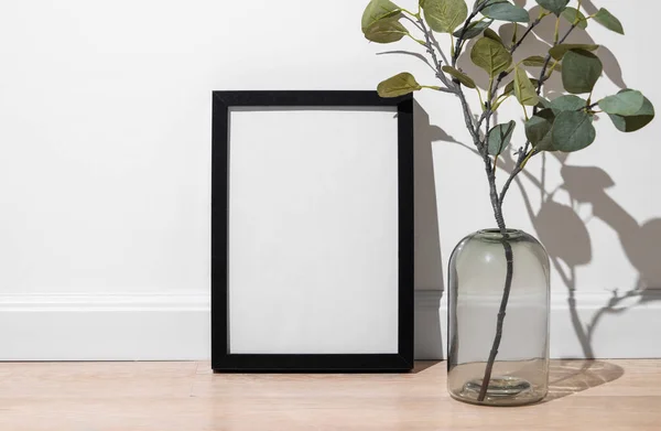 A  black frame with a blank canvas against a light wall and on a wooden floor with vase and branch. Layout concept for photography, poster or painting. Copy space.