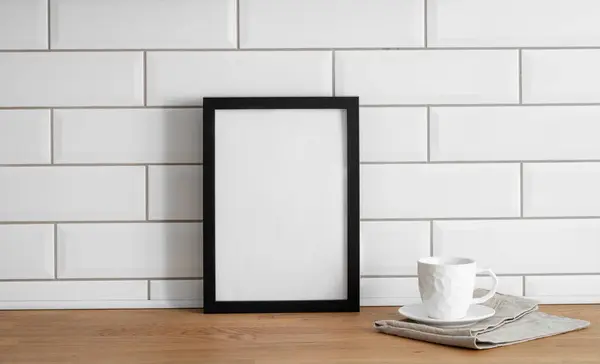 A  frame with a blank canvas against a white tile wall and on a wooden tabletop with cup of tea. Layout concept for photography, poster or painting. Copy space.