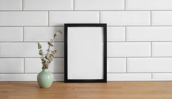 A  frame with a blank canvas against a white tile wall and on a wooden tabletop with vase. Layout concept for photography, poster or painting. Copy space.