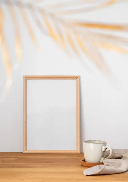 A frame with a blank canvas against a light wall and on a wooden tabletop with blurred palm leaf and cup of tea. Layout concept for photography, poster or painting. Copy space.