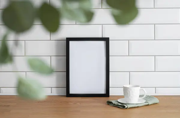 A black frame with a blank canvas against a white tile wall and on a wooden tabletop with blurred green foliage and cup of tea. Layout concept for photography, poster or painting. Copy space.