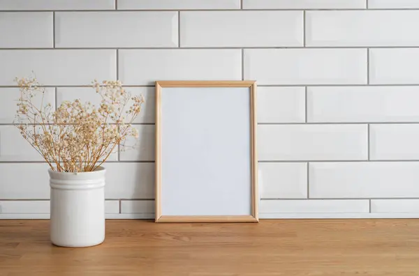 A light frame with a blank canvas against a white tile wall and on a wooden tabletop with dry bouquet. Layout concept for photography, poster or painting. Copy space.