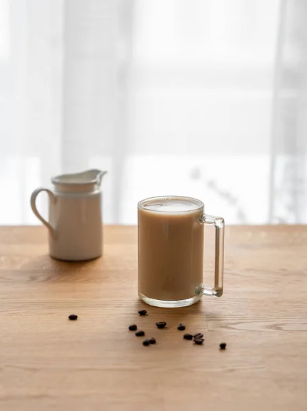 A cup of coffee laltte with milk on a wooden table with coffee beans and milk jug against the background of a window. The concept of a morning tonic drink.
