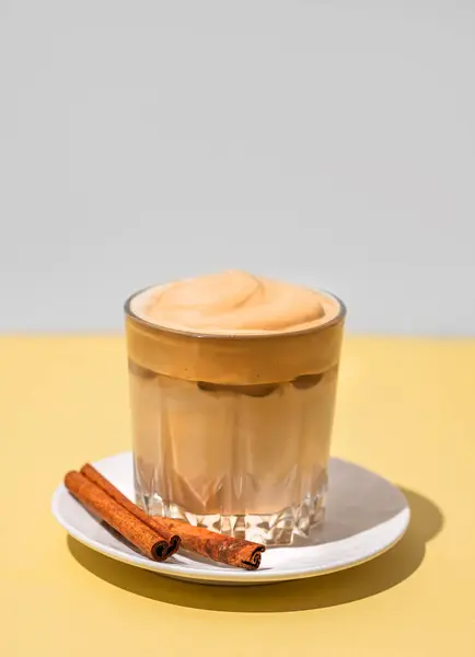Dalgona coffee. Whipped instant iced coffee in a glass with cinnamon sticks on yellow background. The concept of a trendy and popular drink. Free space for text.
