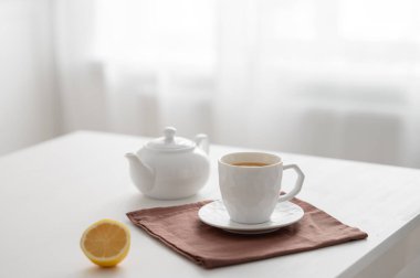 Bir bardak limonlu çay ve mutfak penceresinin arka planında beyaz bir masada bir çaydanlık. Modern İskandinav tarzında iç mekan. Kahvaltı konsepti.