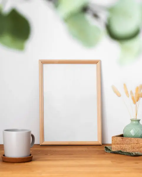 A frame with a blank canvas against a light wall and on a wooden tabletop with blurred green foliage and cup of tea. Layout concept for photography, poster or painting. Copy space.