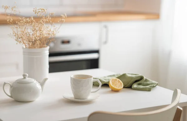 A cup of tea with lemon and a teapot on a white table with dry bouquet. Kitchen with wooden countertop and brick wall in the background. Interior in modern Scandinavian style. Breakfast concept.