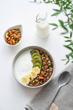 Yeşil dalı ve peçetesi olan beyaz bir masada muz ve kivi ile granola. Düz konsept vejetaryen yoğurt, fındık, müsli ve taze meyve. Üst görünüm. 