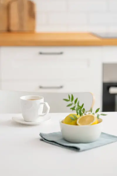 A cup of tea with lemon and a teapot on a white table. Kitchen with wooden empty countertop and brick wall in the background. Interior in modern Scandinavian style. Breakfast concept.