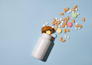 Çeşitli tabletler ve farklı renklerde besin maddeleri açık mavi arkaplandaki beyaz bir şişeden dinamik bir şekilde dökülür. Sağlık hekimi konsepti. Üst görünüm ve kopyalama alanı. 