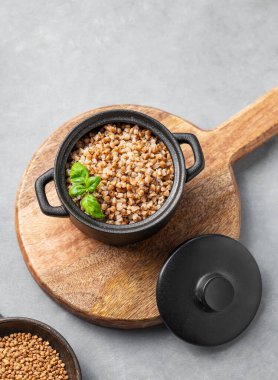 Karabuğday lapası siyah bir tavada, arka planda açık bir zeminde, içinde çiğ taneler olan bir kesme tahtasının üzerinde. Doğal, besleyici ve konforlu yiyecek konsepti. Üst görünüm ve kopyalama alanı.