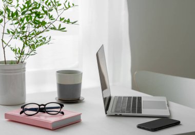 Dizüstü bilgisayarı, kahve fincanı, telefon, not defteri ve güneş ışığı olan bir pencerenin önünde duran gözlükler. İskandinav tarzında rahat ofis kavramı.