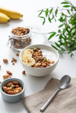 Beyaz ahşap bir masada dallı muz ve yoğurtlu müsli. Doğal ev yapımı diyet kahvaltı konsepti süt ürünü, fındık, müsli ve taze meyveden yapılır..  