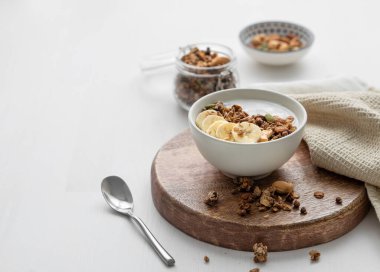 Beyaz bir masanın üzerinde yoğurt ve muz ile pişirilmiş tahıl. Doğal ev yapımı kahvaltı konsepti süt ürünleri, fındık, müsli ve taze meyveden yapılır. Boşluğu kopyala.