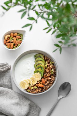 Yeşil dalı olan beyaz bir masada muzlu ve kivili granola. Düz konsept vejetaryen yoğurt, fındık, müsli ve taze meyve. Üst görünüm. 