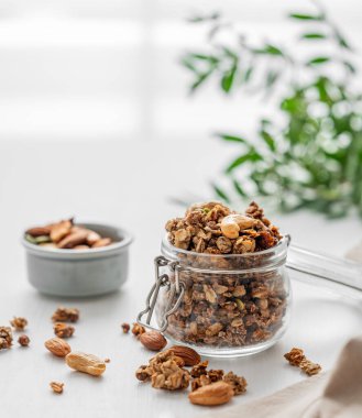 Beyaz arka planda fındıklı, dallı ve pencereli bir kavanozda ev yapımı granola. Rahatlatıcı yemek. Kahvaltıda sağlıklı ve besleyici tahıl müsli konsepti.. 