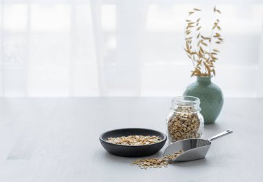 Tabakta yulaf ezmesi, kepçe ve kavanozda mutfak penceresinin önündeki beyaz desenli masada. Sağlıklı bir kahvaltı hazırlamak için doğal vegan mısır gevreği ürünü konsepti. Boşluğu kopyala.