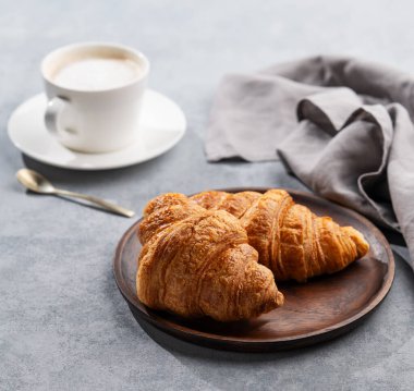 Bir fincan aromatik cappuccino kahve ve sabah ışıklarıyla birlikte ahşap bir tabakta taze kruvasan. Ev yapımı keklerle lezzetli ve sağlıklı bir kahvaltı..