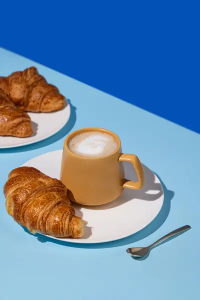 Coffee cup and fresh croissants on blue background with  shadow. Creative layout and concept of healthy food and french breakfast. Copy space.