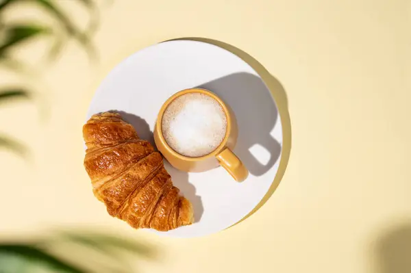 Flat lay of coffee cup and fresh croissant on a white plate on a yellow background with palm leaf shadow. Creative layout and concept of healthy food and french breakfast. Top view and copy space.