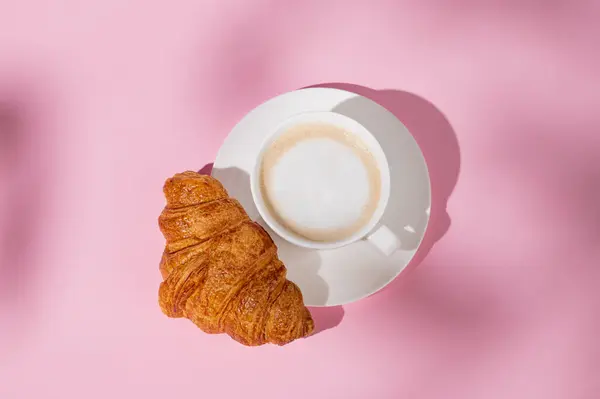 Flat lay of coffee cup and fresh croissant on a white plate on a pink background with shadow. Creative layout and concept of healthy food and french breakfast. Top view and copy space.