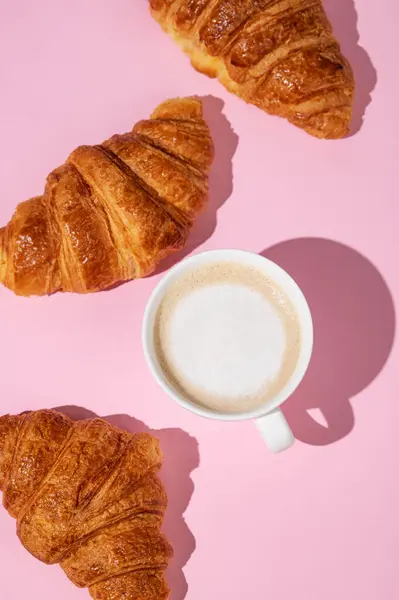 Flat lay of coffee cup and fresh croissants on pink  background with shadow. Creative layout and concept of healthy food and french breakfast. Top view.