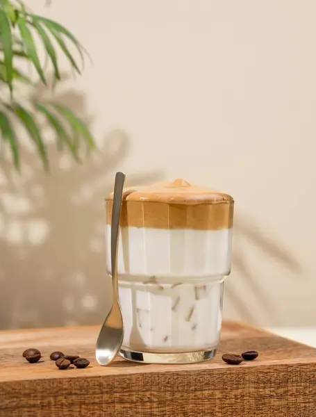 Dalgona coffee. Whipped instant coffee in a glass on a wooden stand with coffee beans on a beige background with palm leaf and shadows. The concept of a trendy and popular drink. Free space for text.
