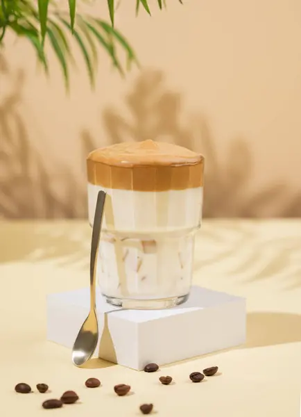 Dalgona coffee. Whipped instant coffee in a glass on a white stand with coffee beans on a beige background with palm leaf and shadows. The concept of a trendy and popular drink. Free space for text.