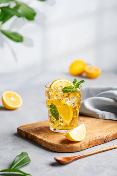 Iced tea with lemon, mint and ice in a glass on a wooden board on a light background with shadow and branch. The concept of a refreshing drink or lemonade on a hot summer day.