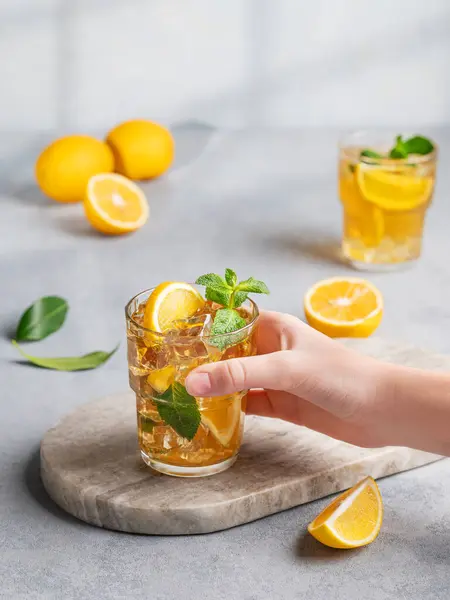 A hand takes a glass of iced tea with lemon and mint against a gray background with citrus fruits and shadows. The concept of a refreshing drink or lemonade on a hot summer day.