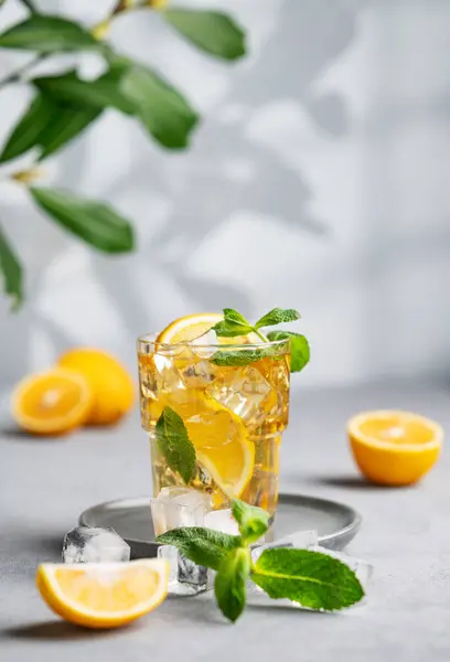 Iced tea with lemon, mint and ice in a glass on a light background with shadow and branch. The concept of a refreshing drink or lemonade on a hot summer day.