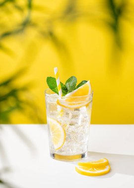 Bir bardak soğuk limonata, nane ve buzlu, sarı ve beyaz arka planda palmiye yapraklı. Sıcak bir yaz gününde ferahlatıcı ve sağlıklı bir içecek konsepti..
