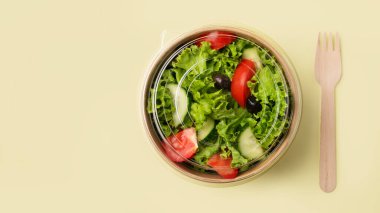 Domatesli taze yeşil salata ve sarı arka planda ahşap çatallı kağıt bir kabın içinde salatalık. Biyolojik olarak ayrıştırılabilir çevre dostu paket servis yemeklerinde sağlıklı gıda konsepti. Üst görünüm.
