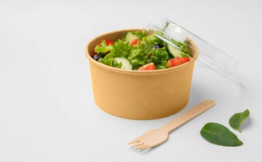 Taze yeşil salata, domatesler ve salatalıklar beyaz arka planda ahşap çatallı kağıt bir kabın içinde. Biyolojik olarak ayrıştırılabilir çevre dostu paket servis yemeklerinde sağlıklı gıda konsepti. Boşluğu kopyala.