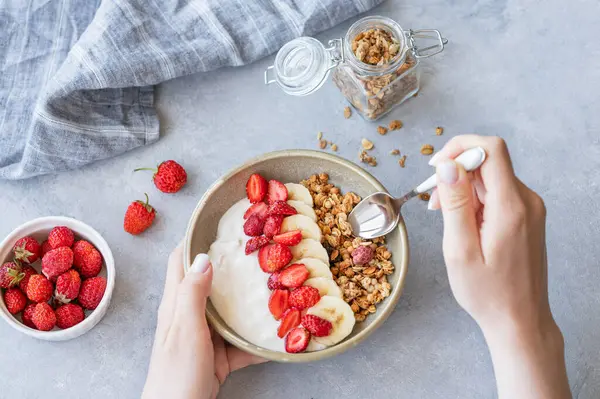 Doğal Yunan yoğurduyla kaşık ve kase tutan kadın eller taze böğürtlen ve peçeteyle mavi zemin üzerinde çilek ve muz. Sağlıklı, besleyici ve besleyici kahvaltı konsepti. Üst görünüm.