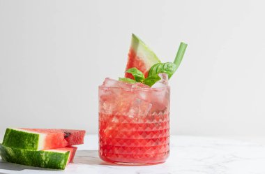 Bir bardak ferahlatıcı karpuz smoothie fesleğenli, beyaz mermer arka planda buz ve bir dilim meyve. Sağlıklı yaz diyeti içeceği ya da kafe ya da bar menüsü için limonata. Ön görünüm, boşluğu kopyala.
