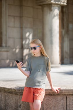 Uzun sarı saçlı, güneş gözlüklü, yeşil tişörtlü ve şortlu genç bir kız tarihi bir binanın önünde duruyor ve elinde bir akıllı telefon tutuyor. 