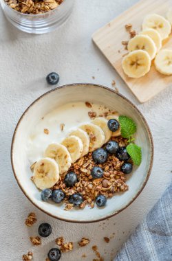 Mısır gevreği ya da müsli ile doğal yoğurt, muz ve yaban mersinleri beyaz arka planda bir kasede sabah ışığında taze böğürtlen. Sağlıklı ve besleyici kahvaltı konsepti. Üst görünüm.