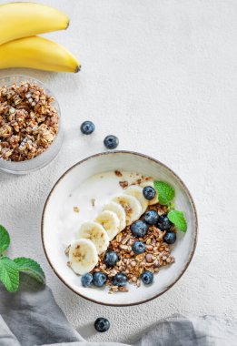 Yoğurt, muz ve yaban mersinli mısır gevreği, arka planda hafif bir kase, peçete ve nane. Süt ürünleri, müsli ve taze meyvelerden sağlıklı kahvaltı kavramı. Üst görünüm 