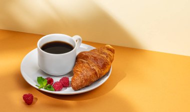 Bir fincan kahve ve gölgeli sarı bir arka planda beyaz bir tabakta ahududu ile taze kruvasan. Sağlıklı yemek ve Fransız kahvaltısı kavramı. Boşluğu kopyala.