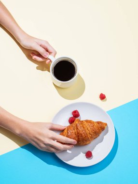 Dişi eller taze kruvasan ve sarı ve mavi arka planda bir fincan espresso kahvesi tutuyor. Kahvaltı pastası modeli ya da afiş için tasarım konsepti. Üst görünüm ve kopyalama alanı.
