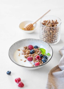 Yoğurtlu granola, ahududu, yabanmersini ve bal beyaz arka planda doğal sabah ışığıyla birlikte bir kasede. Süt ürünleri, yulaf ezmesi ve taze böğürtlenlerden yapılmış ev yapımı kahvaltı konsepti. 