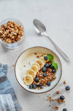 Mısır gevreği ya da müsli, muz ve yabanmersinli Yunan yoğurdu beyaz arka planda bir kasede taze böğürtlen ve sabah ışığında kaşıkla. Sağlıklı ve besleyici kahvaltı konsepti. Üst görünüm 