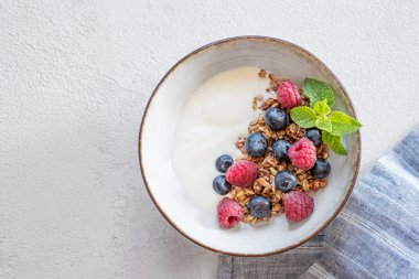 Yunan yoğurdu, ahududu ve yaban mersinli granola beyaz arka planda, peçeteli. Süt ürünlerinin kahvaltısı, sabah ışığında yulaf ezmesi. Üst görünüm ve kopyalama alanı.