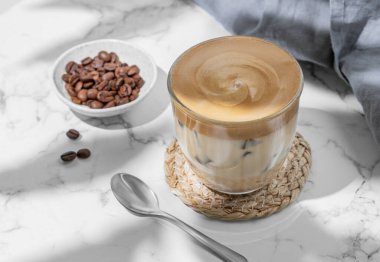 Buzlu dalgona kahvesi. Kahve çekirdekleri, kaşık ve sabah gölgesiyle mermer zemin üzerinde buz olan bir bardakta çırpılmış hazır kahve. Moda ve popüler içki konsepti. Üst görünüm.